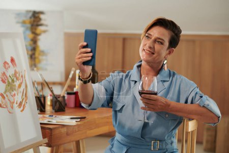Photo for Smiling mature woman taking selfie with glass of wine after finishing picture - Royalty Free Image