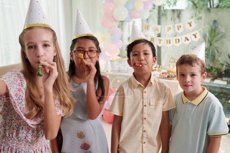 Téléchargez les photos : Groupe d'enfants joyeux soufflant des cornes de fête à la fête d'anniversaire - en image libre de droit