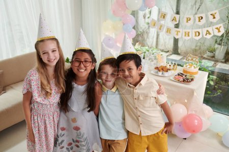 Téléchargez les photos : Joyeux câlin amis dans des chapeaux de fête debout à table avec des friandises et des bonbons - en image libre de droit