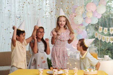 Téléchargez les photos : Groupe d'enfants sautant sous confettis tombant à la fête d'anniversaire - en image libre de droit