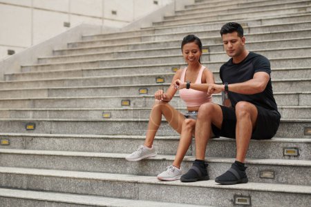 Téléchargez les photos : Couple souriant assis sur les marches et vérifiant les pisteurs de fitness après le jogging du matin - en image libre de droit