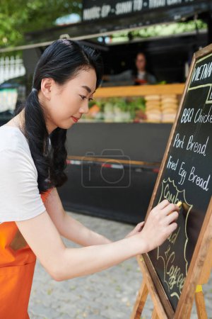 Téléchargez les photos : Souriant jeune femme travaillant pour camion de nourriture de rue écrivant menu - en image libre de droit