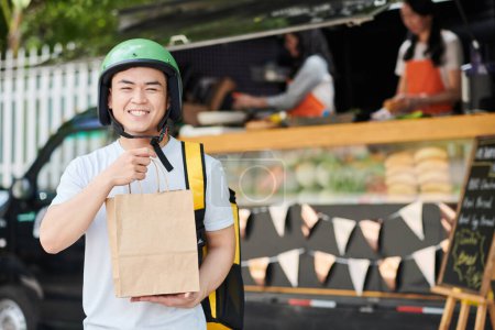 Foto de Retrato de mensajero excitado en casco sosteniendo bolsa de papel con pedido de comida para el cliente - Imagen libre de derechos