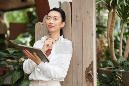 Foto de Retrato de la joven y soñadora empresaria vietnamita con tablet PC apoyada en la puerta de madera y anotando ideas - Imagen libre de derechos