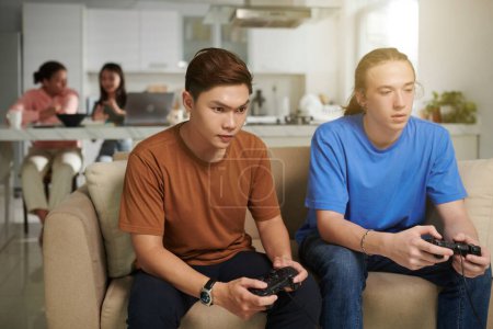 Téléchargez les photos : Adolescents sérieux jouant à des jeux vidéo à la maison lorsque leurs amis font leurs devoirs en arrière-plan - en image libre de droit