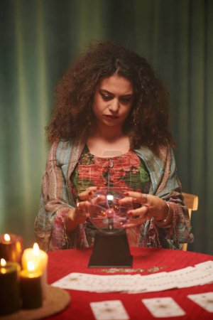 Photo for Portrait of serious witch with poofy hair looking sitting at table with crystal ball - Royalty Free Image