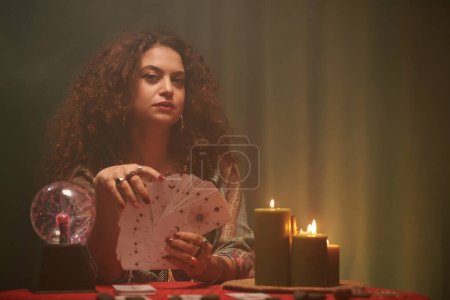 Photo for Portrait of fortune teller with tarot cards sitting at table with burning candles and crystal ball - Royalty Free Image