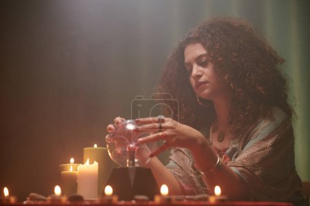 Photo for Pensive sorceress gazing into crystal ball when sitting at table with burning candles - Royalty Free Image