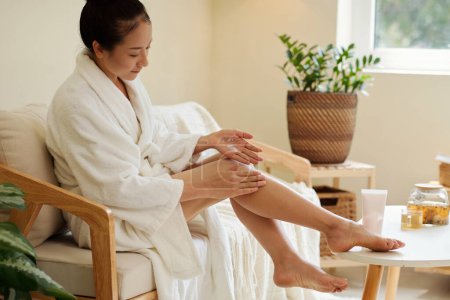 Téléchargez les photos : Jeune femme en peignoir doux appliquant une lotion hydratante sur les jambes après la douche - en image libre de droit