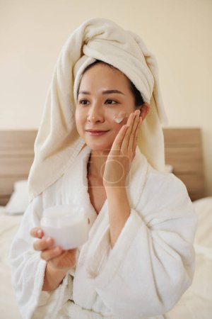 Photo for Smiling young woman applying enriched night face mask after shower - Royalty Free Image