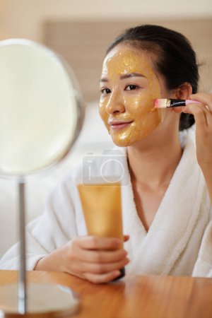 Photo for Happy young woman applying nourishing gel face mask with golden particles - Royalty Free Image