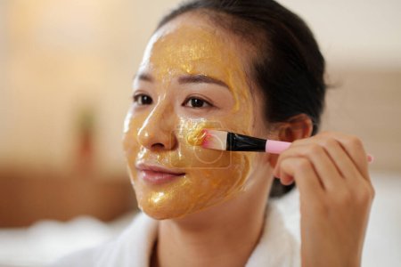 Téléchargez les photos : Jeune femme utilisant un pinceau synthétique lors de l'application d'un masque gel doré pour égayer et revitaliser la peau - en image libre de droit