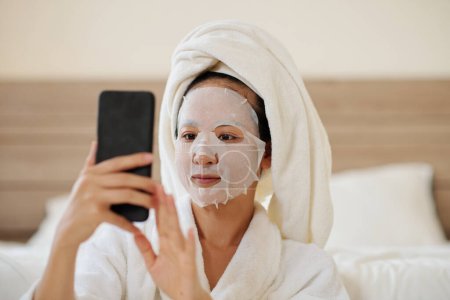 Téléchargez les photos : Jeune femme avec masque de feuille sur le visage textos amis ou vérifier les médias sociaux - en image libre de droit
