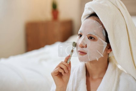 Téléchargez les photos : Jeune femme appliquant un masque en feuille sur le visage et massant avec un rouleau de jade - en image libre de droit