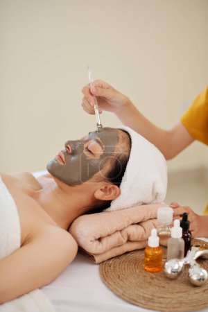 Téléchargez les photos : Cosmetologue application masque d'argile sur le visage de la jeune cliente - en image libre de droit