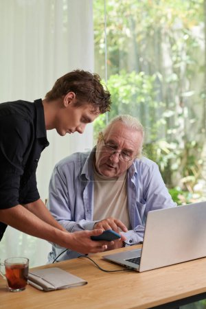 Photo for Teenage boy explaining grandfather how to use mobile application for playing bills and ordering delivery - Royalty Free Image