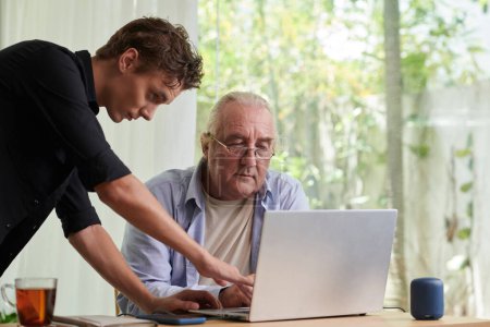 Foto de Hombre joven mostrando padre mayor cómo utilizar el ordenador portátil para la comunicación con la familia - Imagen libre de derechos