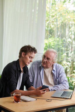 Photo for Senior man asking teenage son to install applications on his smartphone - Royalty Free Image