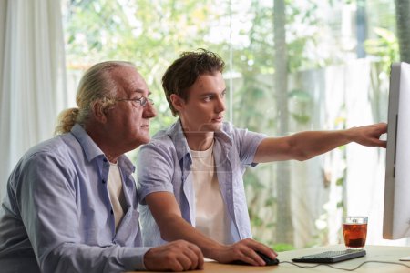 Photo for Teenage boy pointing at laptop screen when explaining grandfather how to use application for online communication - Royalty Free Image