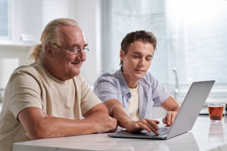 Téléchargez les photos : Jeune homme montrant grand-père âgé comment utiliser diverses applications sur ordinateur portable - en image libre de droit
