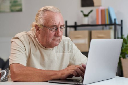 Téléchargez les photos : Homme âgé concentré dans les lunettes de travail sur ordinateur portable à la maison - en image libre de droit