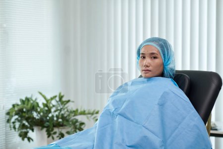Téléchargez les photos : Jeune femme avec des marques sur le visage assis dans la chaise dans le bureau du chirurgien plastique - en image libre de droit