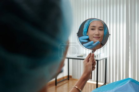 Téléchargez les photos : Heureuse jeune femme asiatique regardant miroir après avoir obtenu un traitement rajeunissant dans la clinique - en image libre de droit