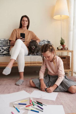Téléchargez les photos : Fille dessin sur le sol à la maison lorsque sa mère et son petit chien reposant sur le canapé en arrière-plan - en image libre de droit