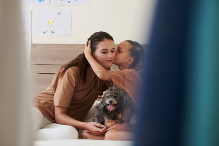 Photo for Girl kissing mother on cheek when they sitting on bed with small dog - Royalty Free Image