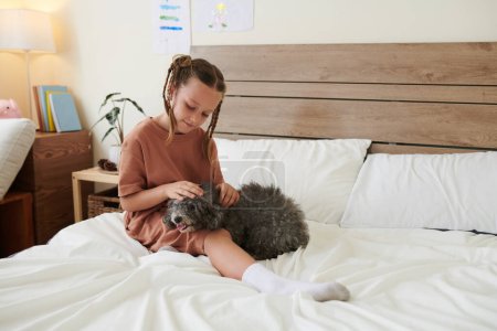 Photo for Smiling girl with braided hair sitting on bed in her room and patting dog - Royalty Free Image