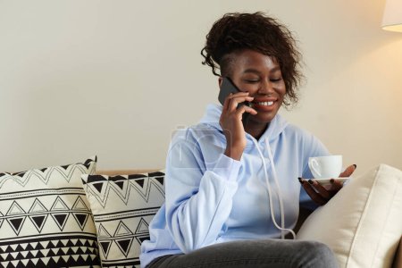 Foto de Retrato de una joven negra positiva con una taza de café en la mano hablando por teléfono con un amigo - Imagen libre de derechos