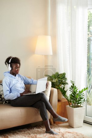 Téléchargez les photos : Souriant jeune femme dans casque assis sur counch à la maison et regarder l'épisode de l'émission sur l'écran de l'ordinateur portable - en image libre de droit
