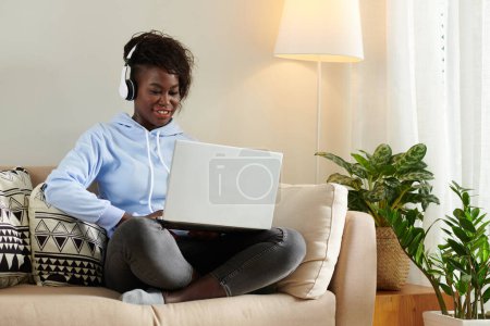 Téléchargez les photos : Positif jeune femme noire portant un casque lorsque vous jouez à un jeu sur ordinateur portable à la maison - en image libre de droit