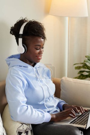 Foto de Sonriente joven mujer negra escuchando música en los auriculares cuando se trabaja en el ordenador portátil en casa - Imagen libre de derechos