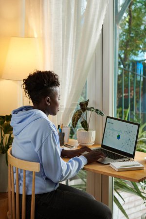 Téléchargez les photos : Concentré jeune femme noire analyser les données dans le rapport sur ordinateur portable lorsque vous travaillez à la maison - en image libre de droit