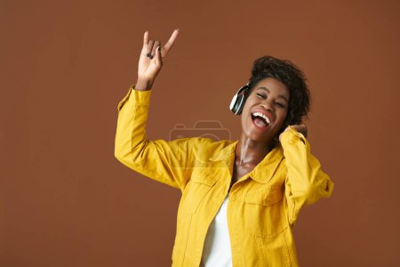 Foto de Mujer joven y feliz escuchando música rock en auriculares, aislada en marrón - Imagen libre de derechos