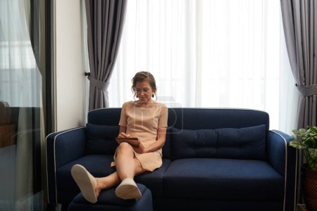 Foto de Mujer de mediana edad concentrada sentada en el sofá y contrato de revisión en la tableta - Imagen libre de derechos