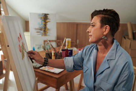 Téléchargez les photos : Souriant créatif femme mature peinture fleurs sur chevalet dans son atelier - en image libre de droit