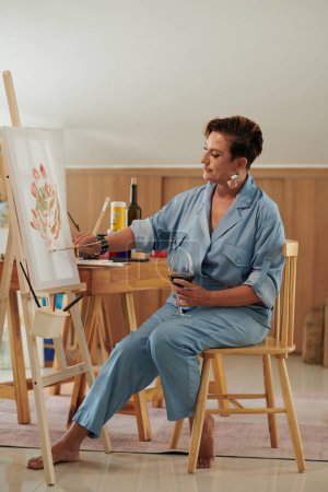 Foto de Mujer de mediana edad bebiendo vino y pintando en caballete en casa - Imagen libre de derechos