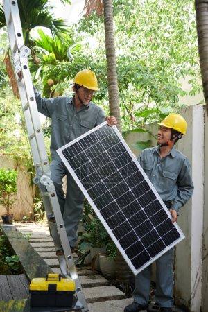 Foto de Contratista ayudando a su compañero de trabajo a levantar el panel solar en el techo - Imagen libre de derechos