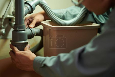 Téléchargez les photos : Image rapprochée du travailleur des services à domicile nettoyant un tuyau en plastique sous un évier de cuisine - en image libre de droit