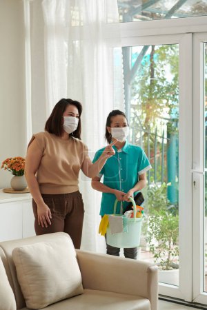 Photo for Woman in protective mask talking to cleaning service worker with bucket of tools and detergents - Royalty Free Image
