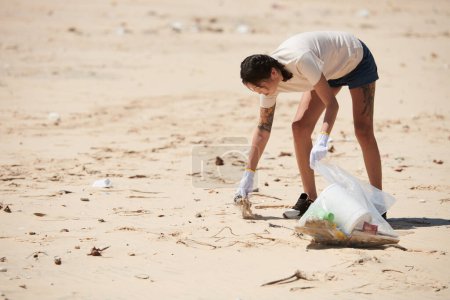 Téléchargez les photos : Jeune femme portant des gants textiles lors de la collecte des ordures sur la plage de sable le jour de la Terre - en image libre de droit