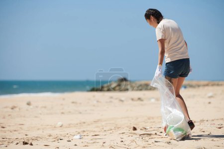 Photo for Young woman walking on beach with big plastic bag and collecting trash brought by sea wave - Royalty Free Image