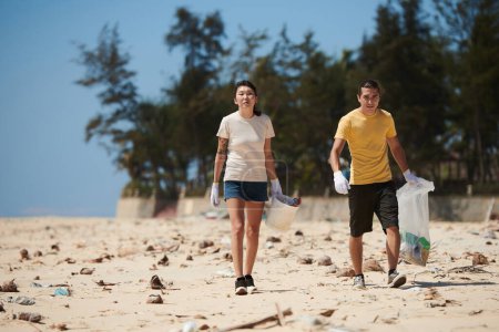 Téléchargez les photos : Jeune couple marchant sur une plage de sable et ramassant des ordures sur une plage sale - en image libre de droit