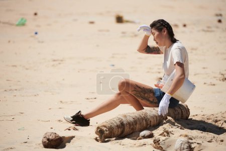 Téléchargez les photos : Fatiguée jeune femme assise sur un camion de palmiers après avoir ramassé des ordures sur la plage le jour de la Terre - en image libre de droit