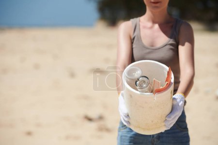 Téléchargez les photos : Image recadrée d'une femme vounteer montrant un seau avec des bouteilles en verre et des déchets en plastique qu'elle a trouvés sur une plage de sable fin - en image libre de droit