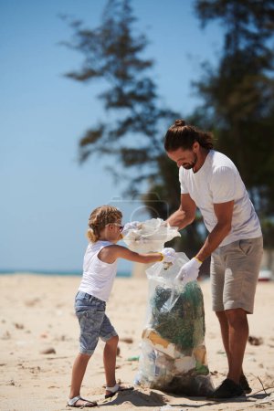Téléchargez les photos : Petite fille aidant père à mettre les ordures qu'ils ont trouvé sur la plage dans un grand sac en plastique - en image libre de droit