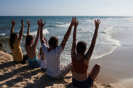 Foto de Grupo de jóvenes emocionados sentados en la playa de arena, levantando aram y mirando las olas - Imagen libre de derechos