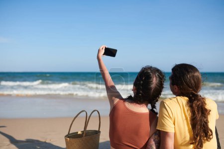 Téléchargez les photos : Jeune femme prenant selfie avec petit ami en se reposant sur la plage, vue de dos - en image libre de droit